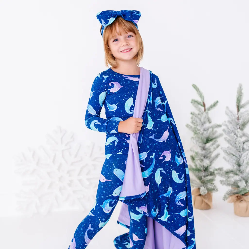 A child in matching pajamas smiling at the camera, a royal blue blanket with a lavender reverse in a print of blue and lavender narwhals draped over one arm.